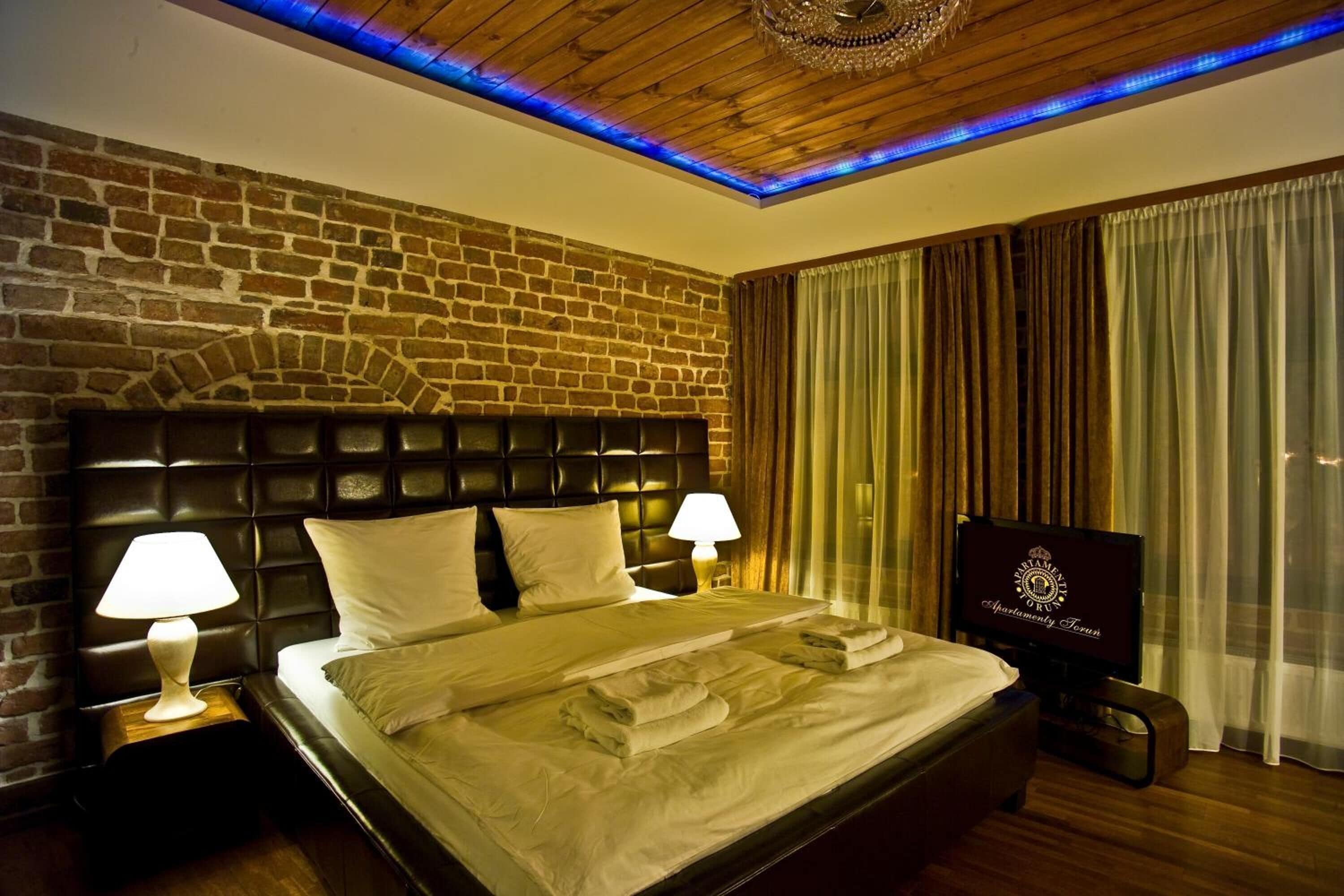A hotel room with a brick wall, a wooden ceiling, a bed with white linens, two bedside lamps, and a television on a stand.