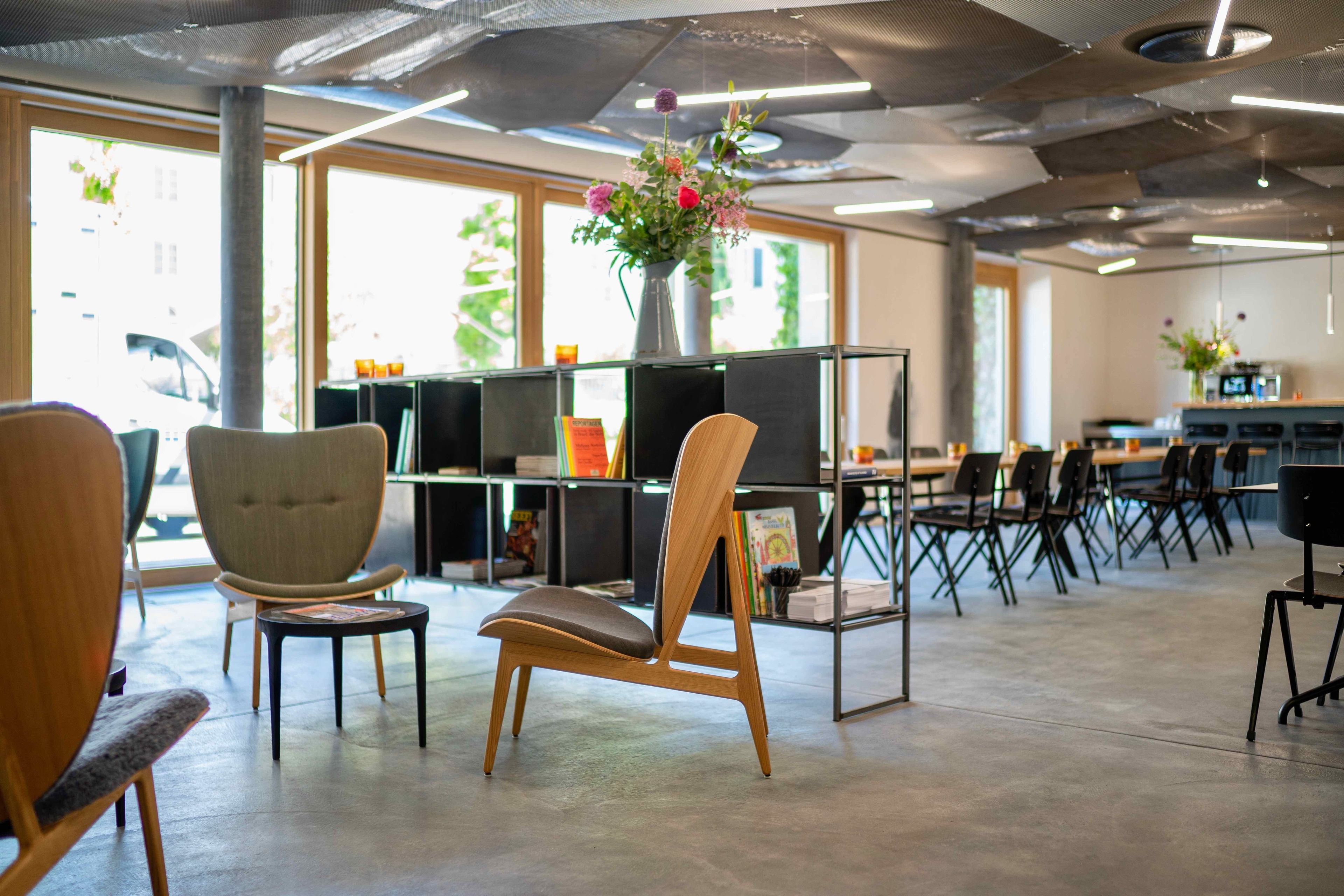 A modern library or reading room with wooden chairs, a central table, and a bookshelf with books and a vase of flowers.