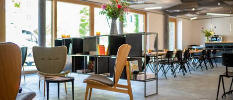A modern library or reading room with wooden chairs, a central table, and a bookshelf with books and a vase of flowers.