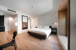 Un dormitorio moderno con suelo de madera, una cama con ropa blanca, una mesita de noche pequeña y una lámpara de pared.