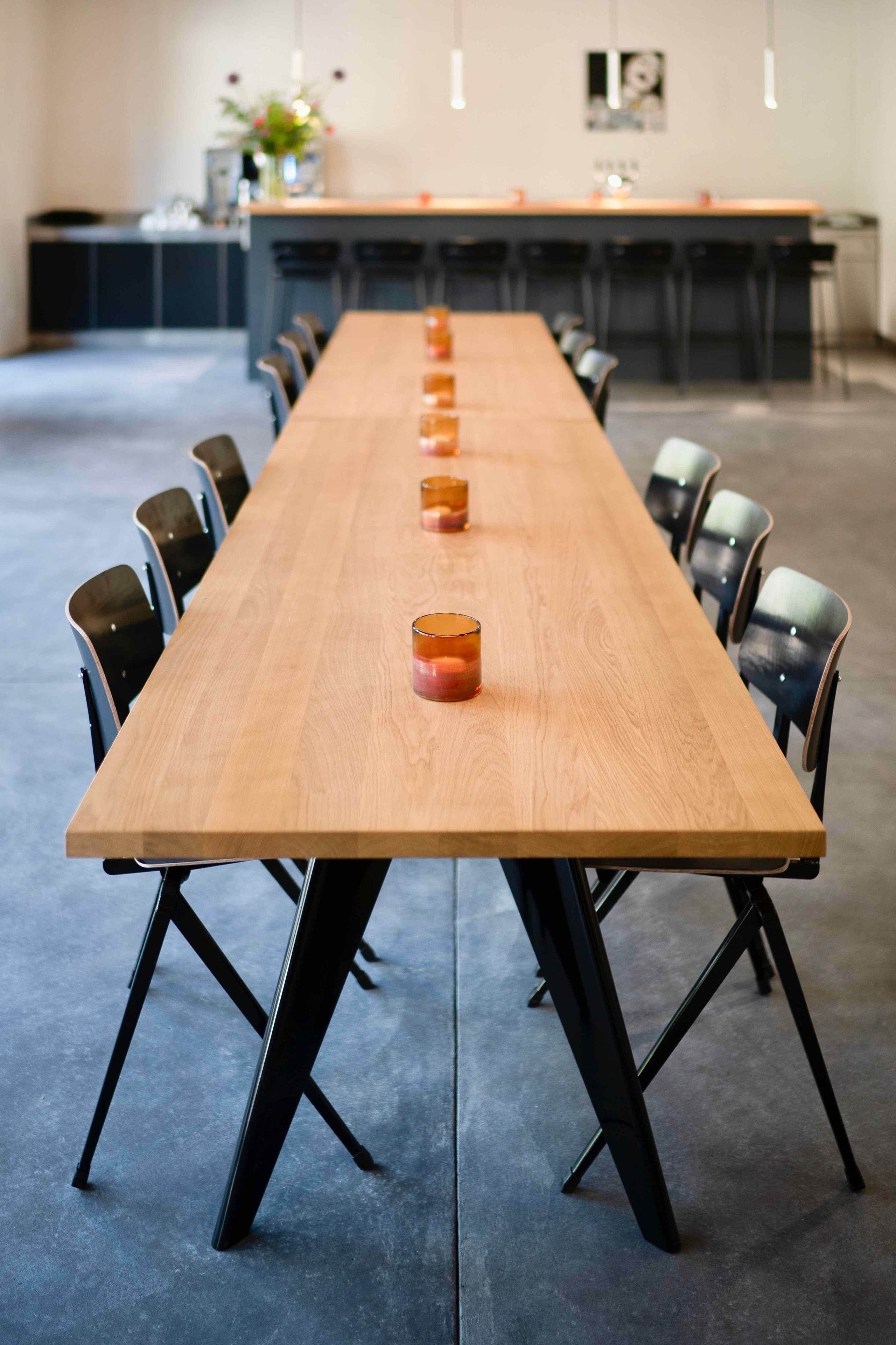 Un lungo tavolo da conferenza in legno con sedie nere, un'area bar sullo sfondo e una cucina moderna visibile attraverso una porta aperta.