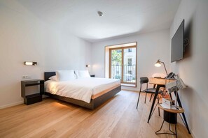 Un dormitorio moderno con cama, mesitas de noche, un escritorio con silla, televisor y ventana con vista a edificios.
