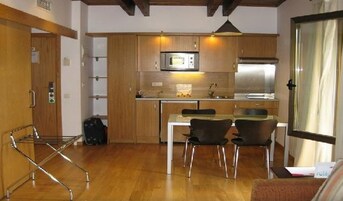 A compact kitchen with wooden cabinets, a microwave, and a stainless steel refrigerator. There is a dining table with two chairs and a small window with curtains.