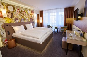 A hotel room with a large bed, a desk with a chair, a small table with a vase, and a wall with a decorative pattern. - Das Schokoladenhotel (Westerstede)