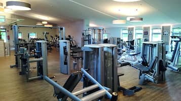 Une salle de sport équipée de divers appareils d’exercice, dotée d’un plancher en bois et de grands luminaires au plafond.