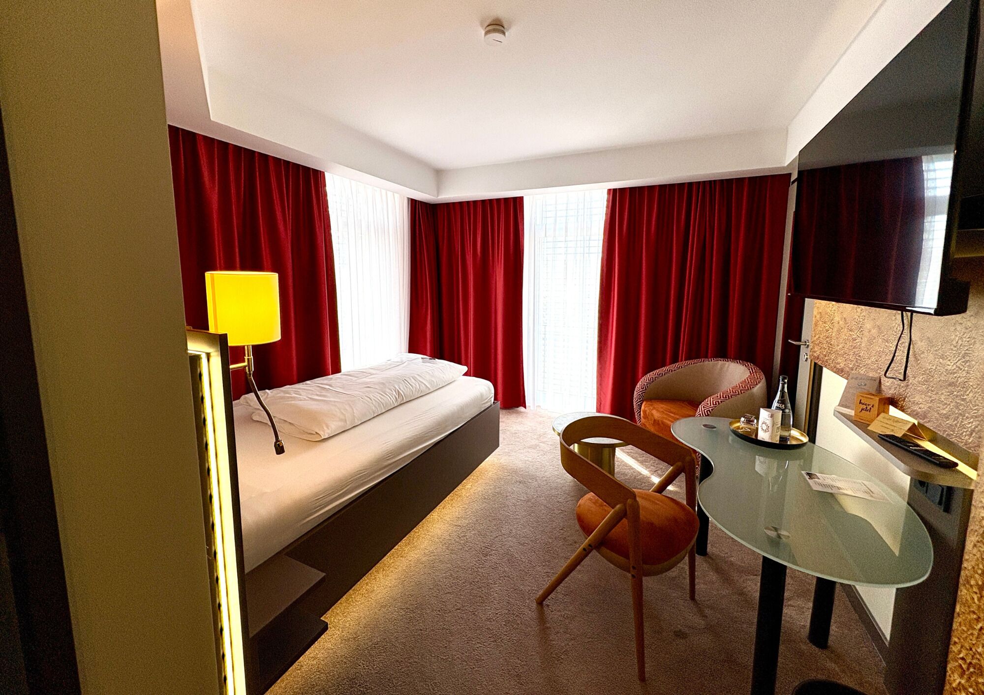 Une chambre d’hôtel avec un lit, un bureau et une chaise, une télévision et des rideaux rouges.