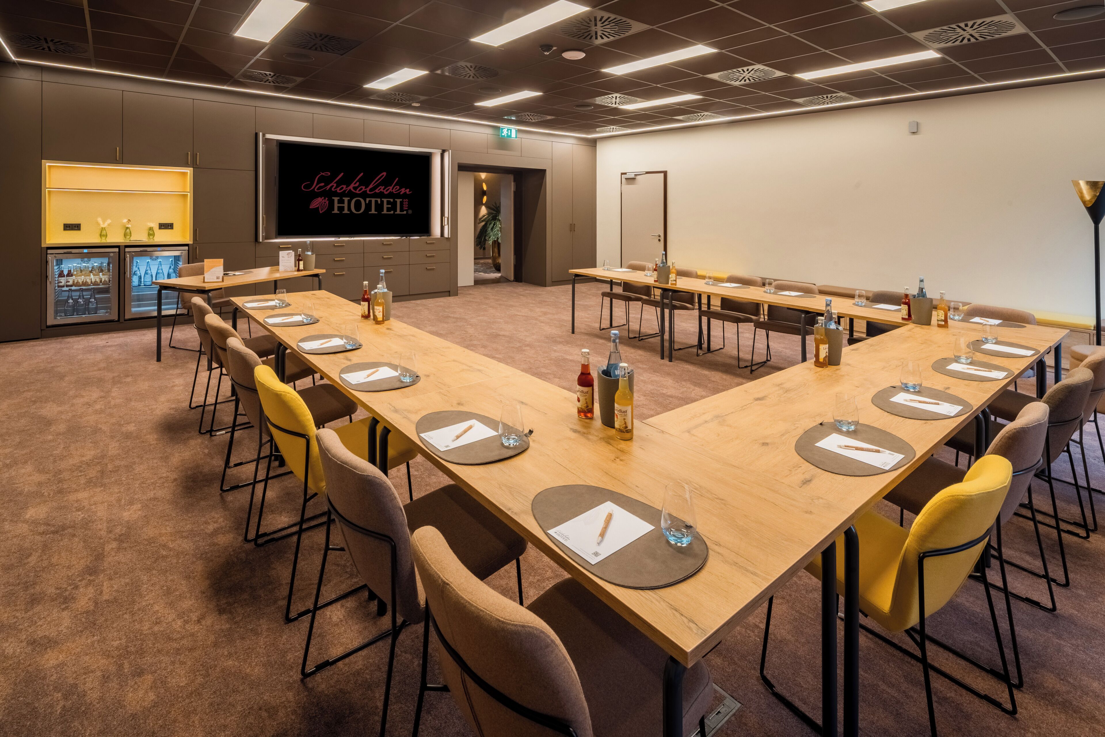 Ein Konferenzraum mit langen Tischen, Stühlen und einem großen Bildschirm, der 'Schokaden HOTEL' anzeigt.