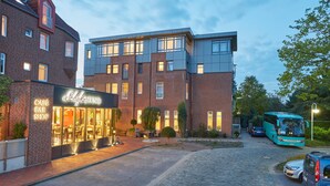 A building with a cafe and bar shop on the ground floor, a modern brick structure with large windows, and a parked bus in the foreground. - Das Schokoladenhotel (Westerstede)