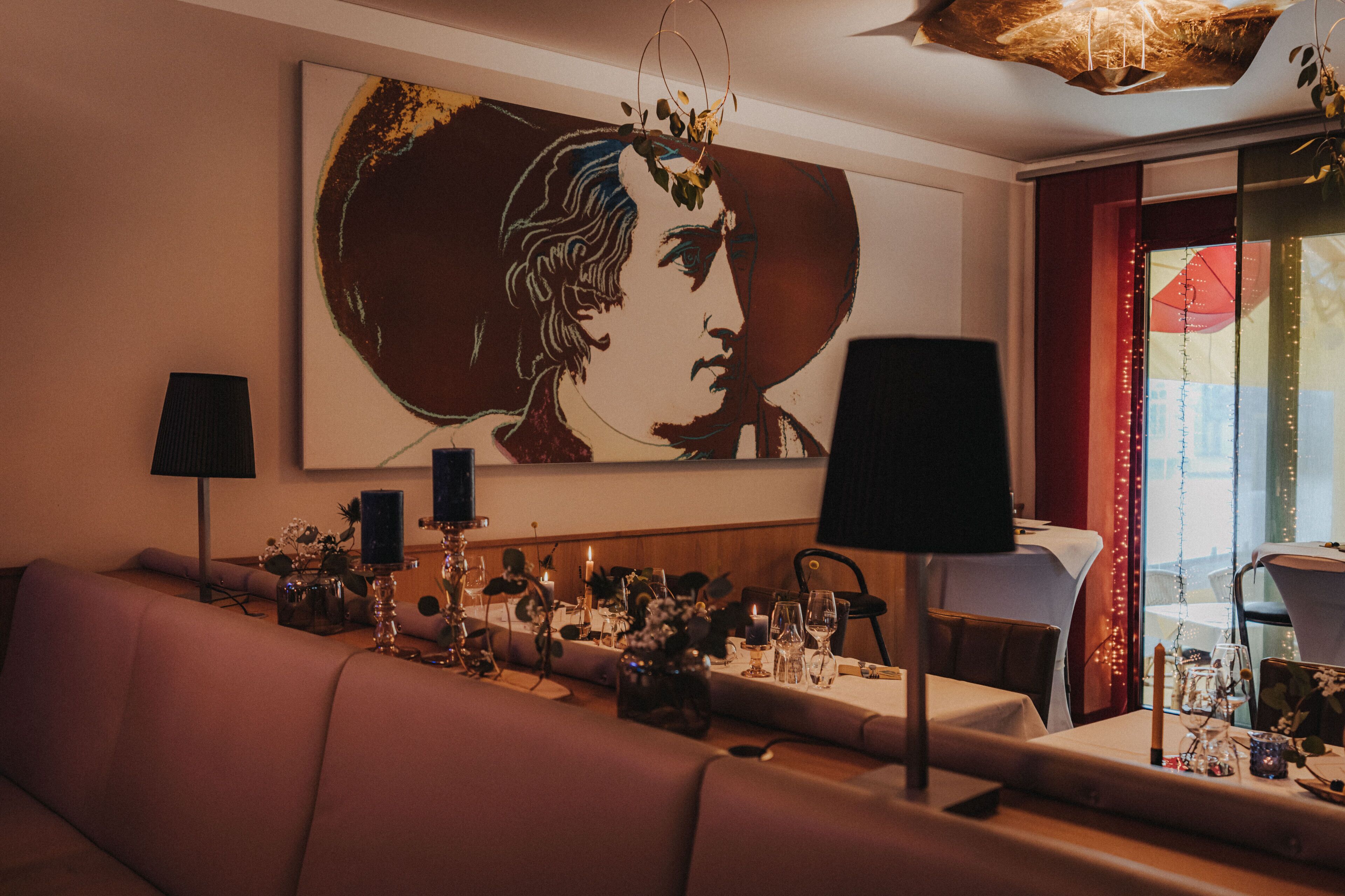 A dining area with a long table set for a meal, a sofa, and a large painting on the wall.