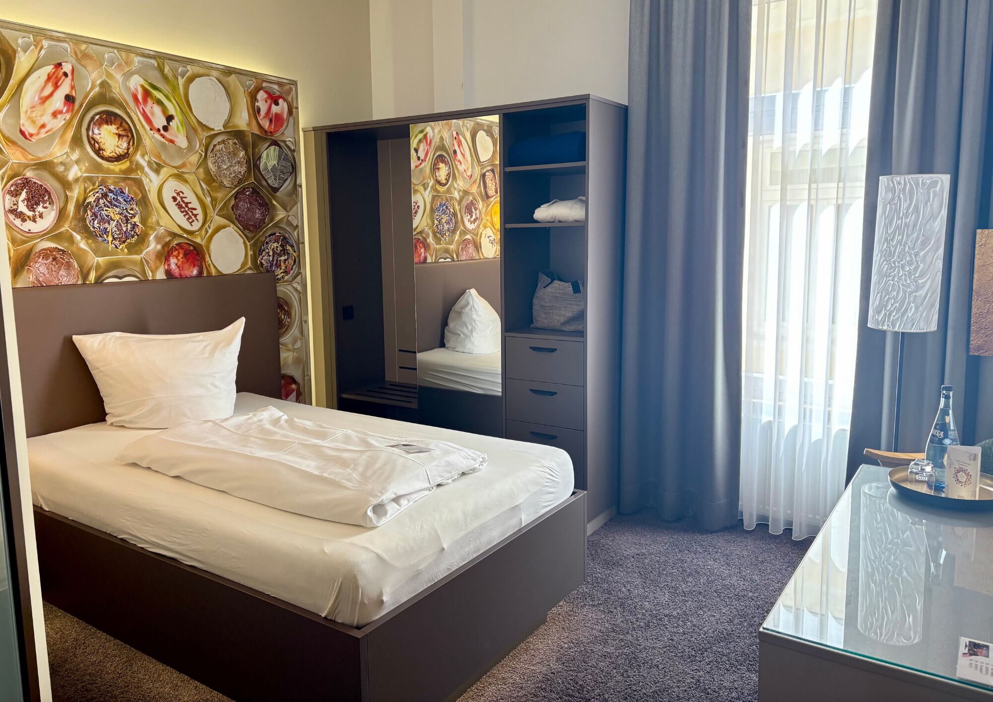 A hotel room with a bed, a wardrobe, a desk with a lamp, and a large window with curtains.
