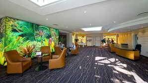 A hotel lobby with a large digital rainforest mural, comfortable seating, a reception desk, and a patterned carpet. - Das Schokoladenhotel (Westerstede)