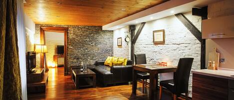 A cozy living room with a stone wall, wooden flooring, a sofa with a yellow pillow, a dining table with chairs, and a kitchen area with wooden cabinets.