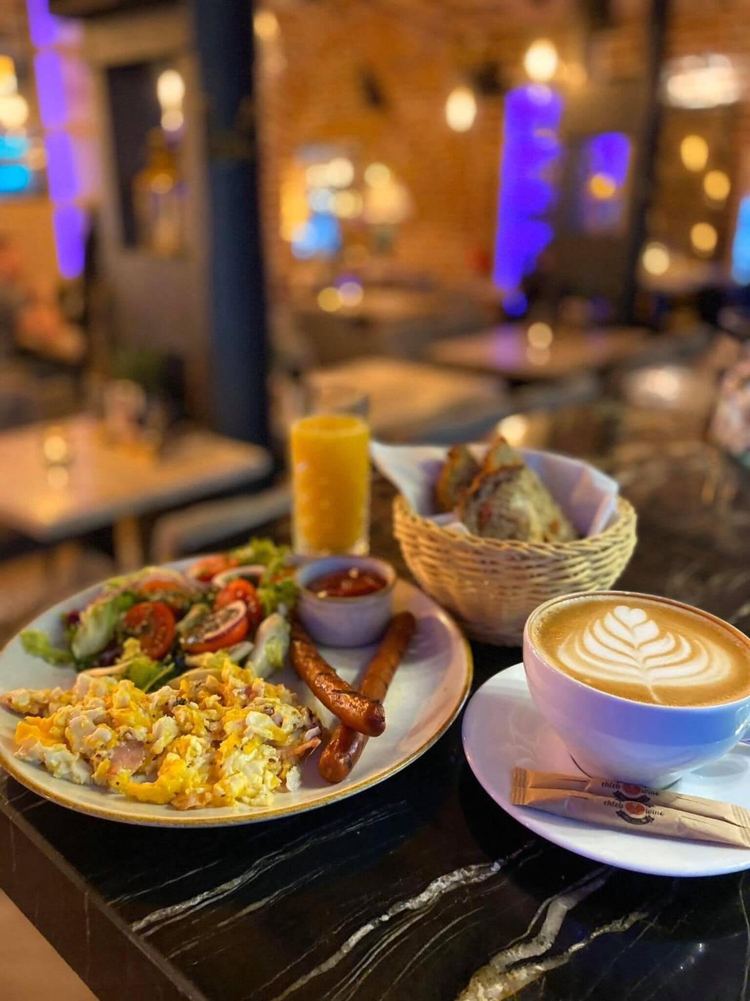A breakfast setting with a plate of scrambled eggs, sausages, and a salad, a cup of coffee with latte art, and a basket of bread.