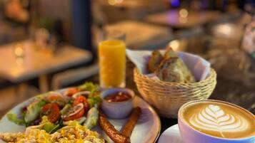 Un desayuno con una fuente de huevos revueltos, salchichas y una ensalada, una taza de café con arte de latte, y una cesta de pan.