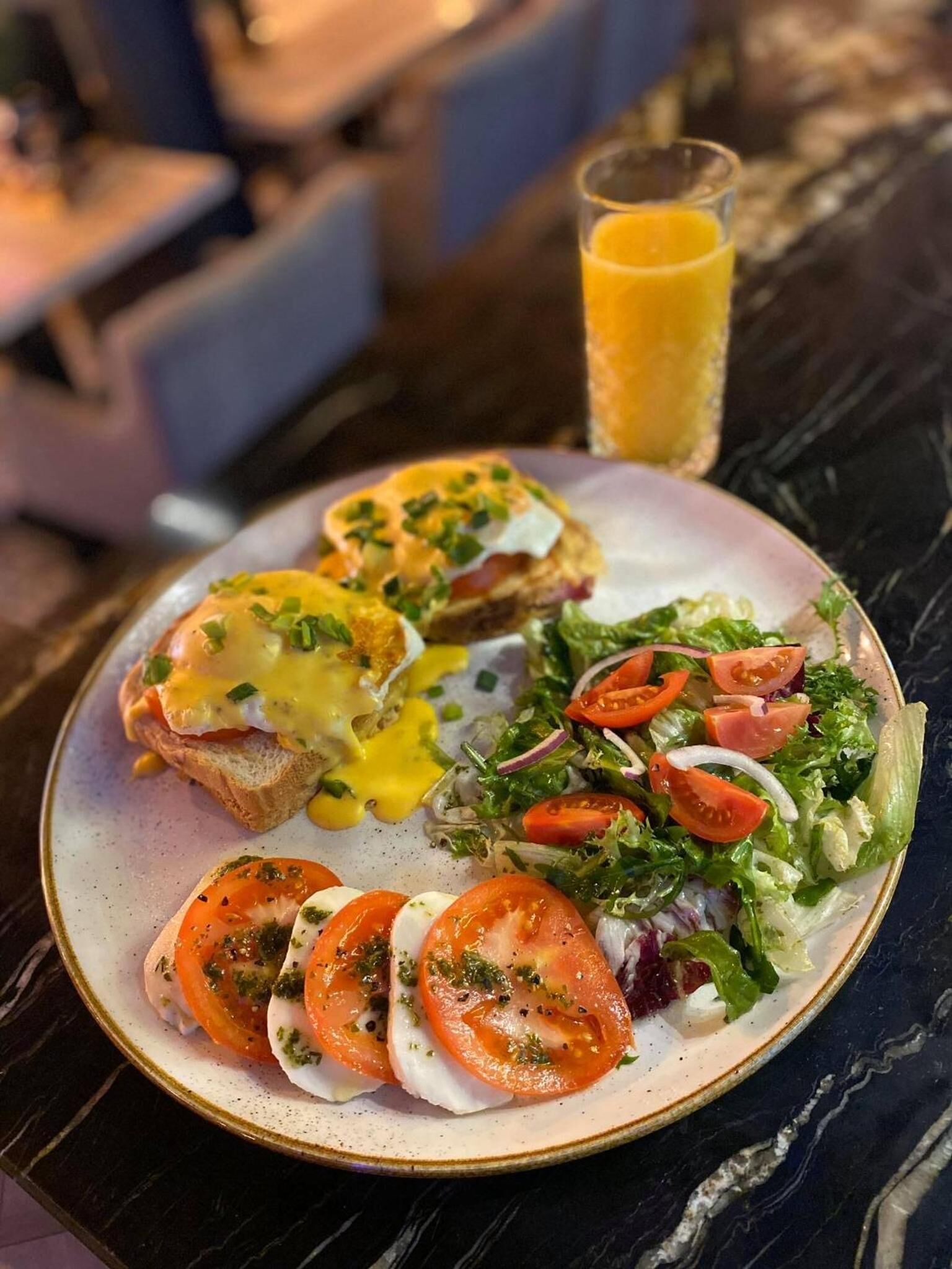 Un plato de desayuno con huevos Benedictinos, ensalada y un vaso de jugo de naranja sobre una mesa de mármol.