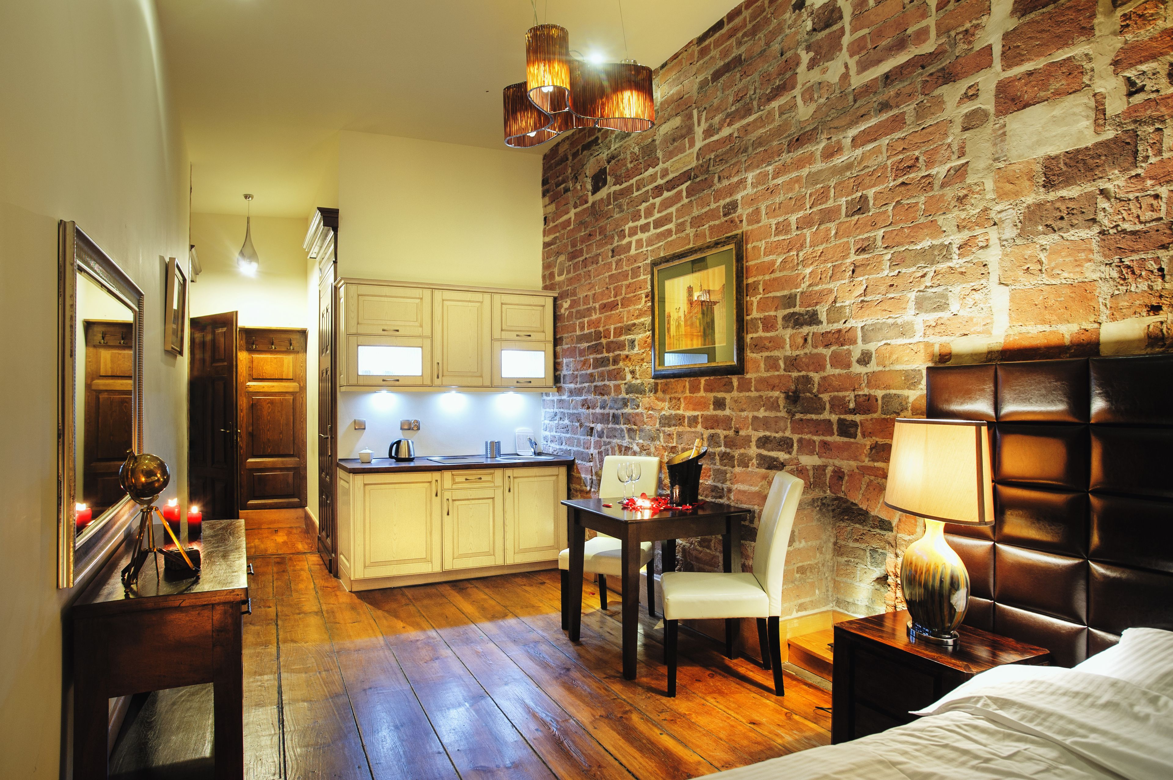 Una habitación con pared de ladrillo, suelo de madera, una mesa de comedor con sillas, una cama y una mesita de noche con lámpara.
