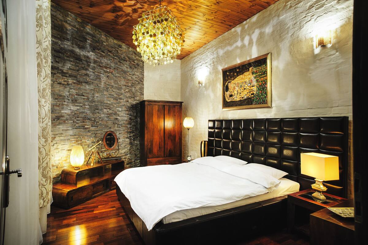 A bedroom with a wooden ceiling, a stone wall, a bed with a black headboard, and a chandelier.