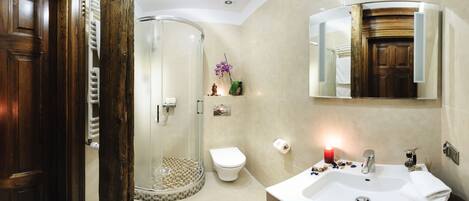 Un baño con puerta de madera, cabina de ducha de vidrio, inodoro, lavamanos con espejo y una vela roja en la encimera.