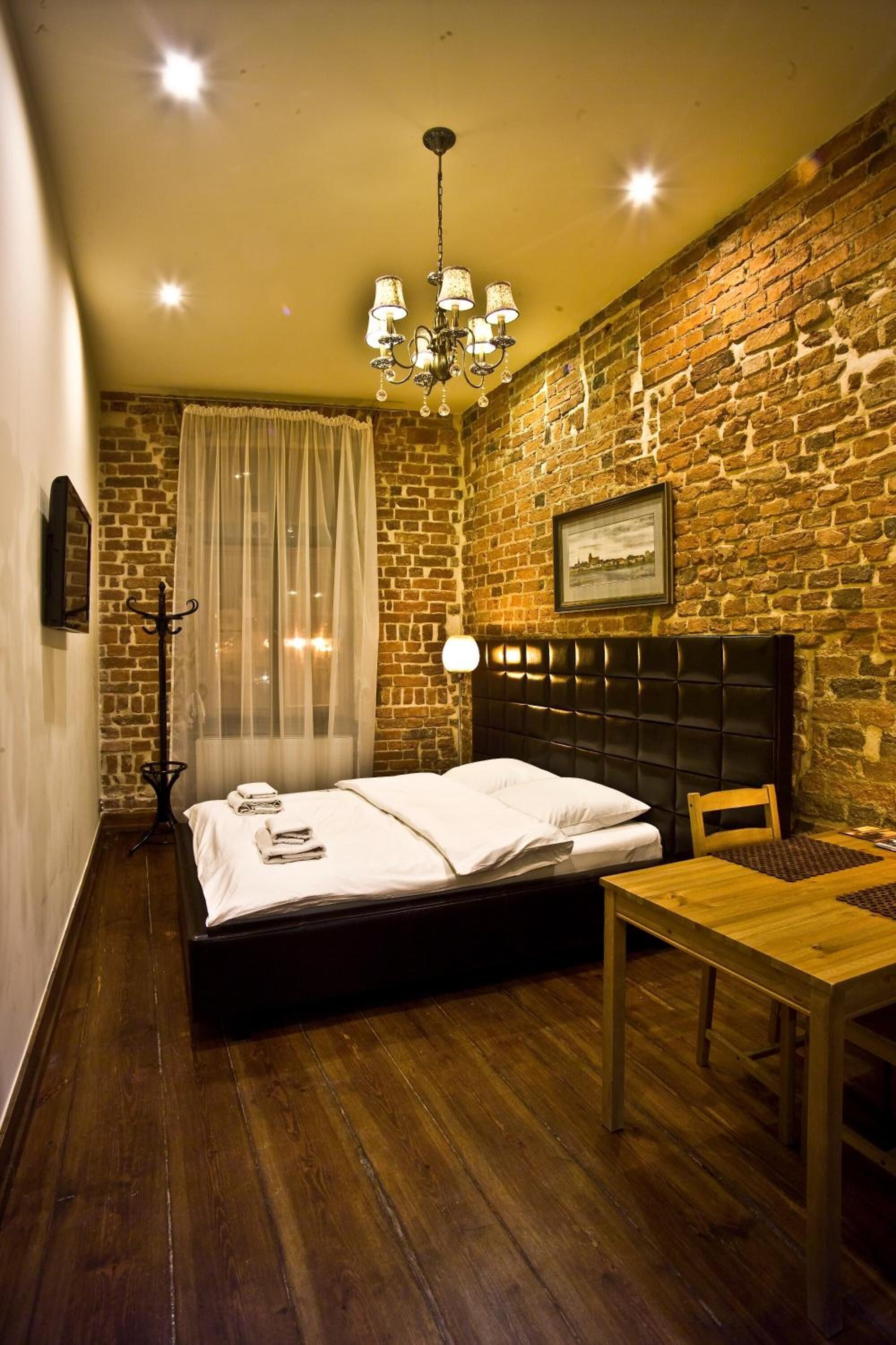 Quarto com parede de tijolos, piso de madeira, cama com roupa de cama branca, mesa de madeira, lustre e cabideiro.