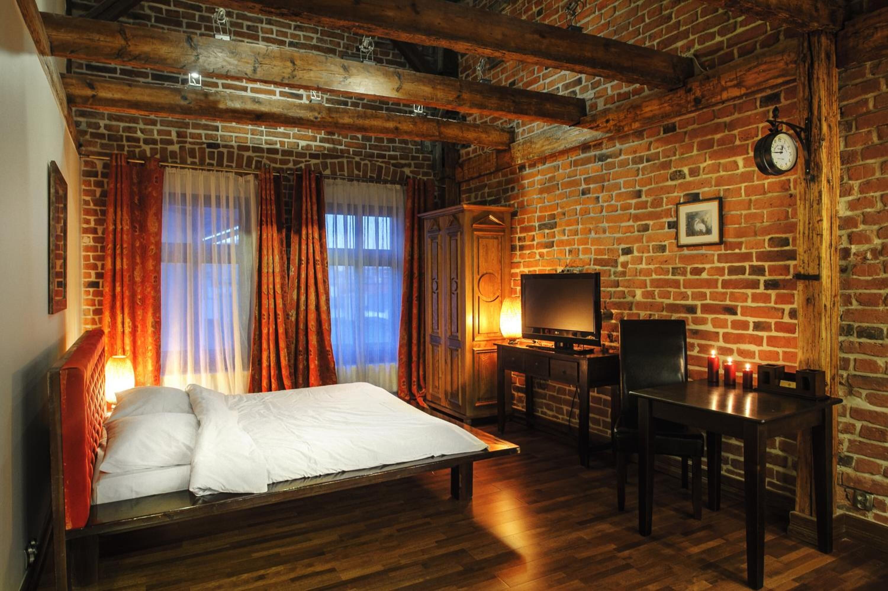 A room with a brick wall, wooden beams, a bed, a desk, and a television.