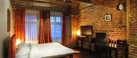 Quarto com parede de tijolos, vigas de madeira, cama, escrivaninha e televisão.