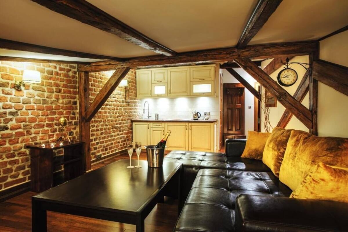 A cozy room with a brick wall, wooden beams, a dining table with glasses, a sofa, and a kitchen in the background.