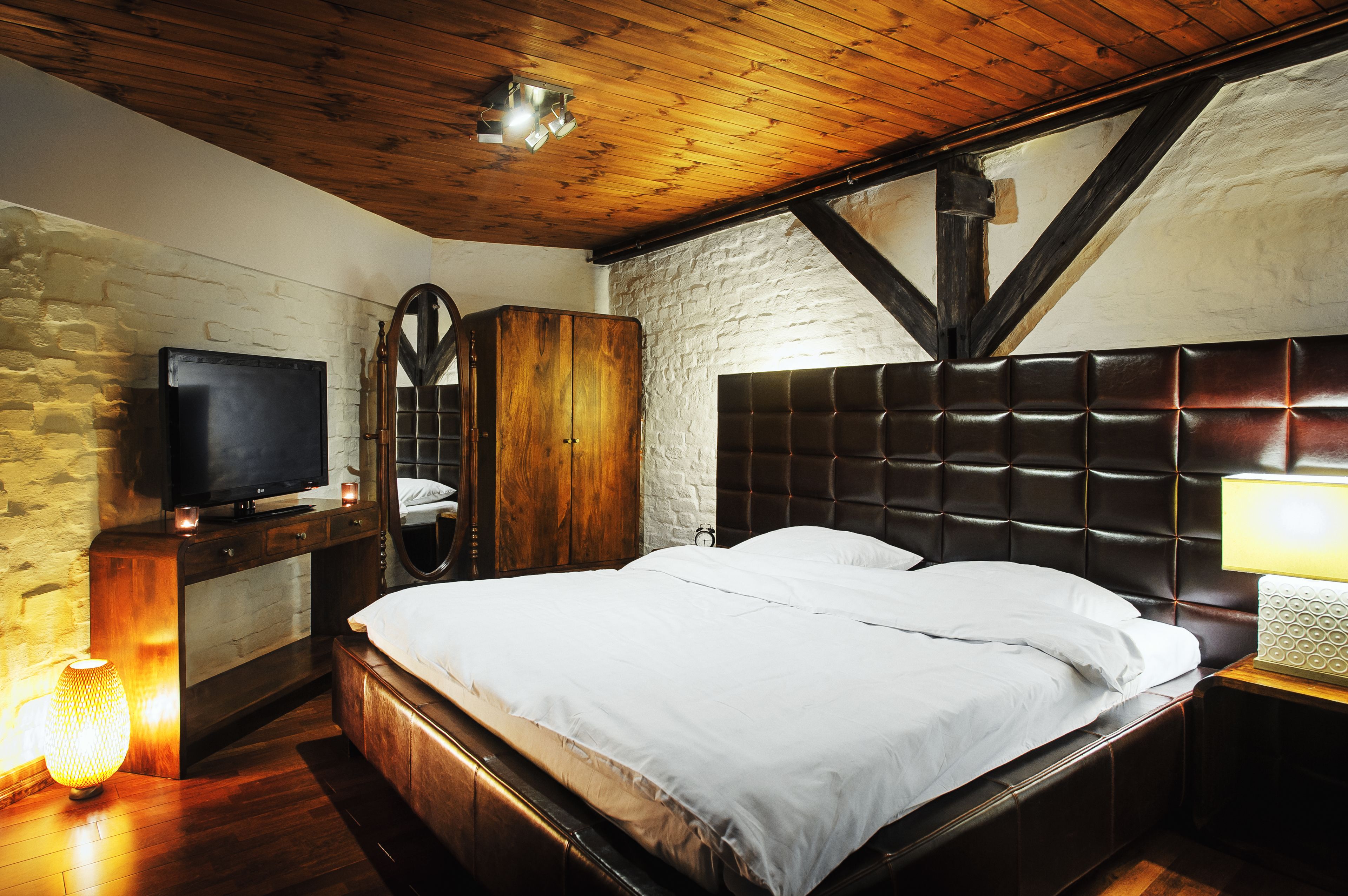 Un dormitorio con techo de madera, pared de piedra, televisor, armario, cama con ropa de cama blanca y una lámpara de noche.