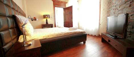 Un dormitorio con cama, lámpara de noche, suelo de madera, pared de ladrillo y televisión sobre un mueble.