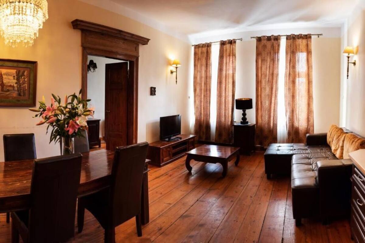 A room with a wooden dining table, chairs, a sofa, a TV, and a chandelier.