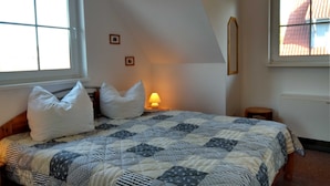 Ein Schlafzimmer mit einem Bett, Kissen, einer Nachttischlampe, einem kleinen hölzernen Hocker und einem Fenster.