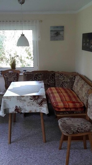 Un coin repas avec une table et des chaises, un canapé agrémenté d’un coussin à carreaux, une horloge au mur et une plante en pot près de la fenêtre.