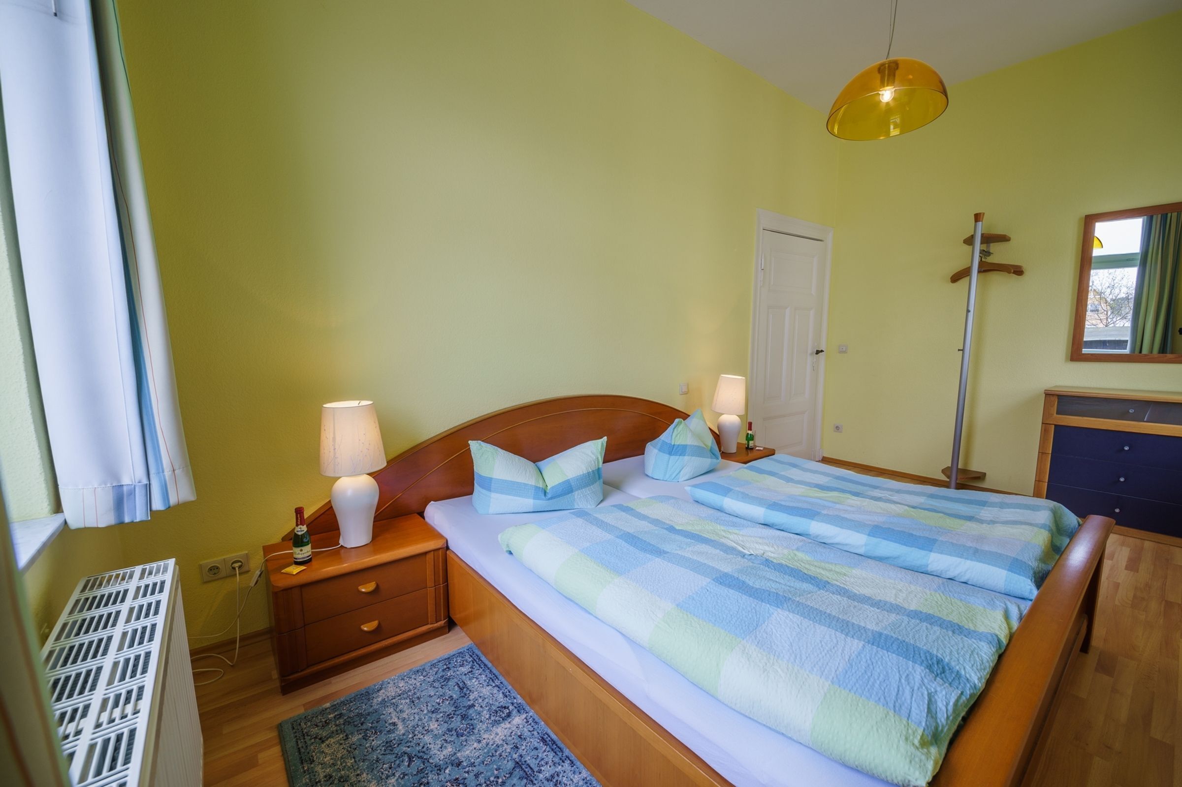 Une chambre à coucher avec un lit en bois, deux lampes de chevet, une table de nuit, un radiateur, un tapis bleu et un luminaire suspendu.