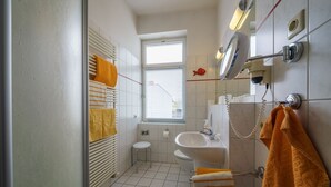 Ein Badezimmer mit Toilette, Waschbecken und Spiegel. Ein Fenster mit Jalousien, ein Handtuchhalter und ein kleiner Hocker sind ebenfalls vorhanden.