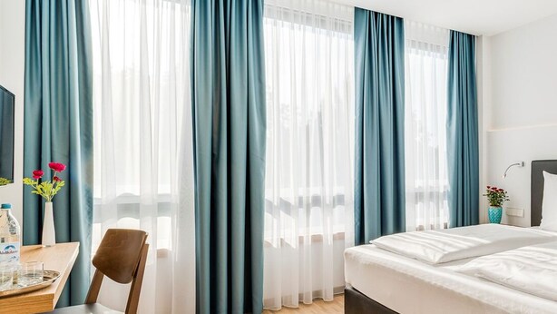 A hotel room with a bed, a desk with a chair, a vase with flowers, and curtains.