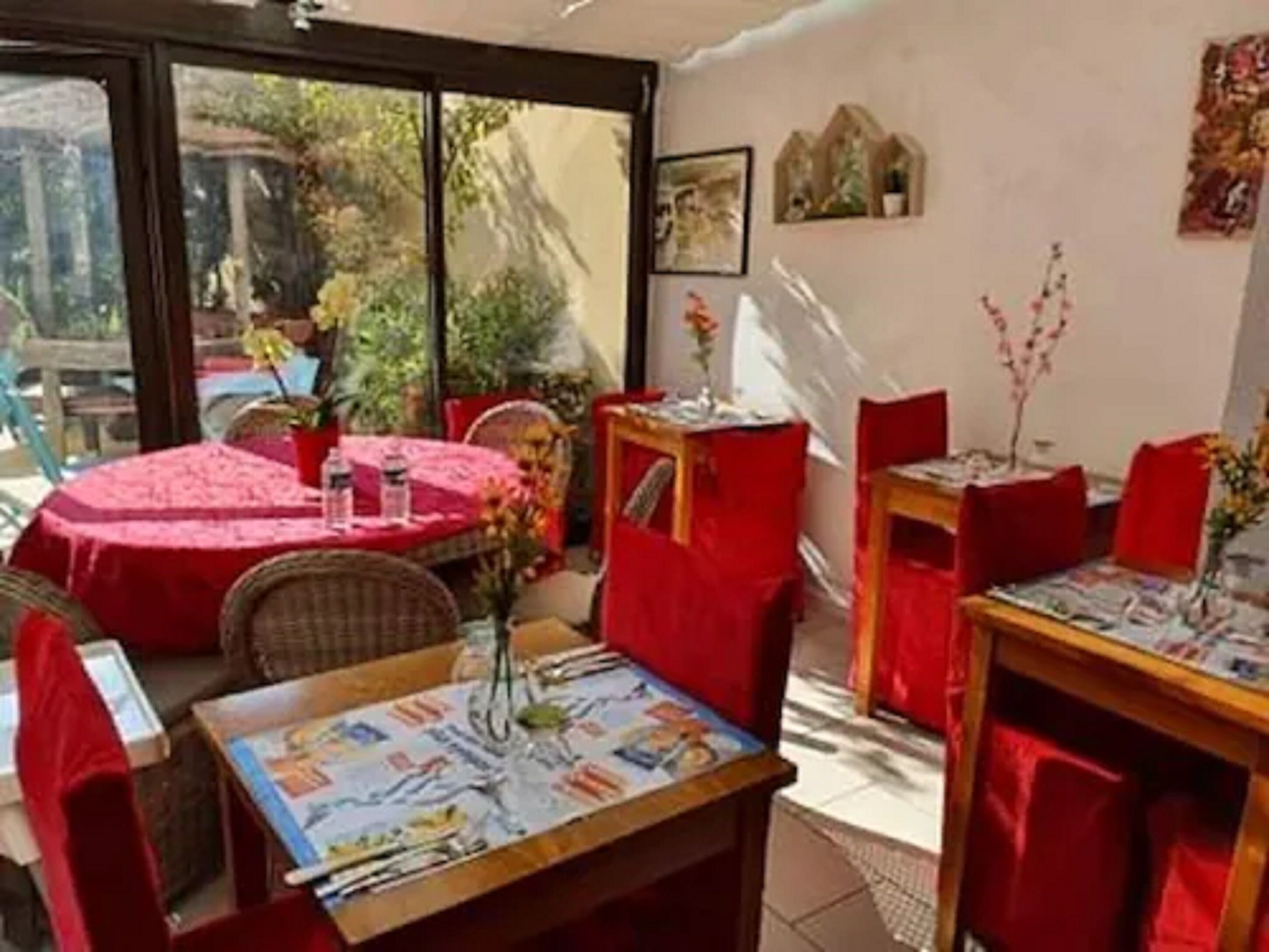 A dining area with red tablecloths, wicker chairs, and a view of greenery outside.