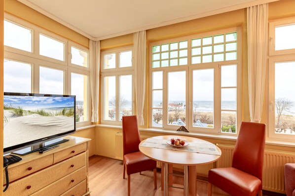 A room with a TV, a wooden table with chairs, and a view of the beach through large windows.