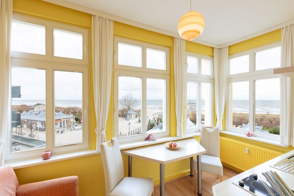 A room with a view of the beach, a table with chairs, and a window with curtains.