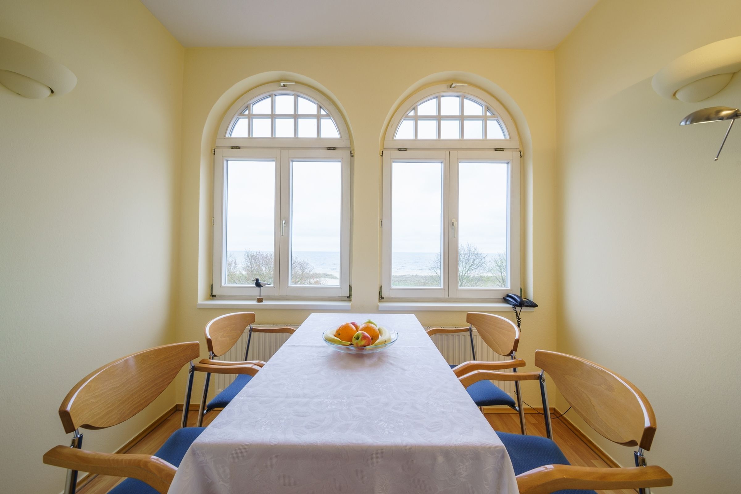 A dining room with a table set for four, two windows with arched tops, and a bowl of fruit on the table.