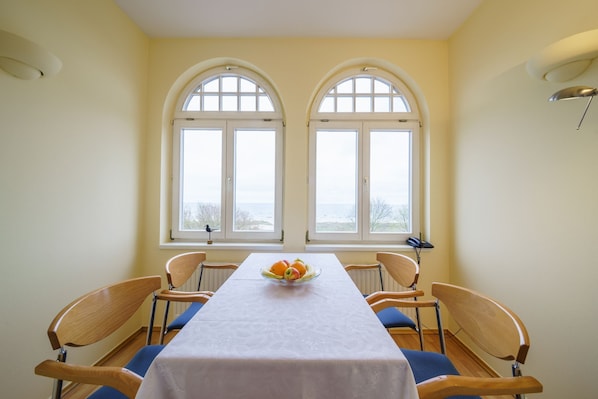 Une salle à manger avec une table dressée pour quatre personnes, deux fenêtres surmontées d’arcs, et un saladier de fruits posé sur la table.