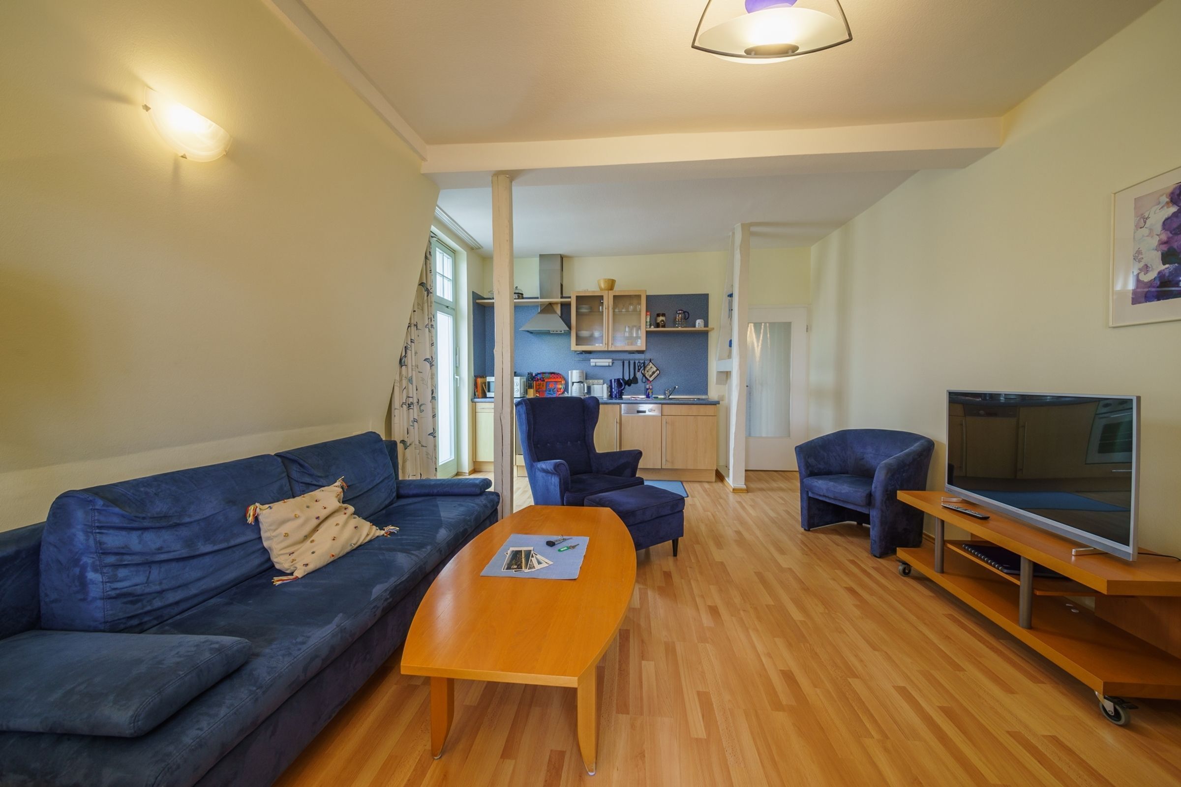 Una sala de estar con sofá, reposapiés y mesa de centro. Hay un televisor sobre un mueble bajo y una cocina al fondo.