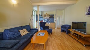A living room with a sofa, ottoman, and coffee table. There is a TV on a stand and a kitchen in the background. - Vacation apartment 6 - Villa HENRIETTE (Heringsdorf)