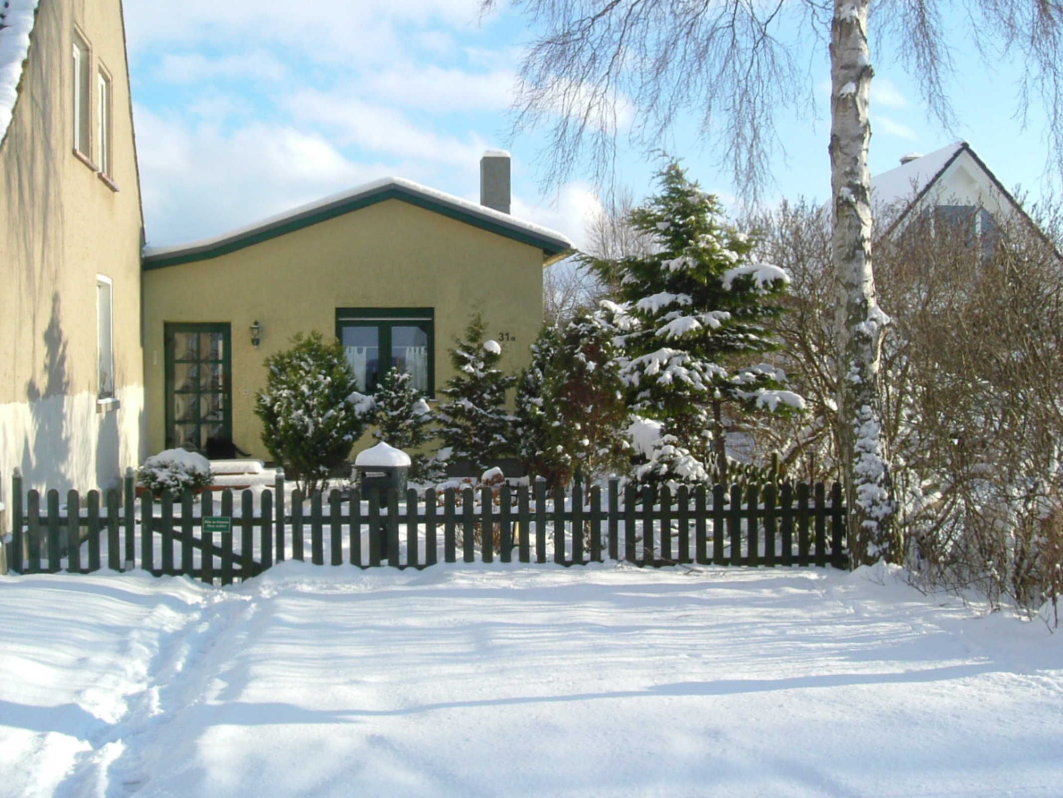 A snow-covered house with a green door, a picket fence, and a snow-covered tree in front.