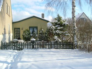 Una casa cubierta de nieve con una puerta verde, una valla de piquetes y un árbol nevado al frente.