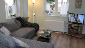 A living room with a sofa, a glass coffee table, a TV on a stand, a radiator, and a window with curtains. - Apartment Bodden - View of the water - Apartments Polchow - directly at the (Glowe)