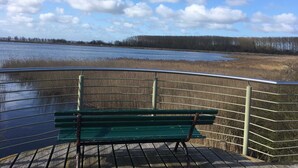 A bench on a wooden deck overlooking a body of water with a grassy bank and trees in the background. - Apartment Bodden - View of the water - Apartments Polchow - directly at the (Glowe)