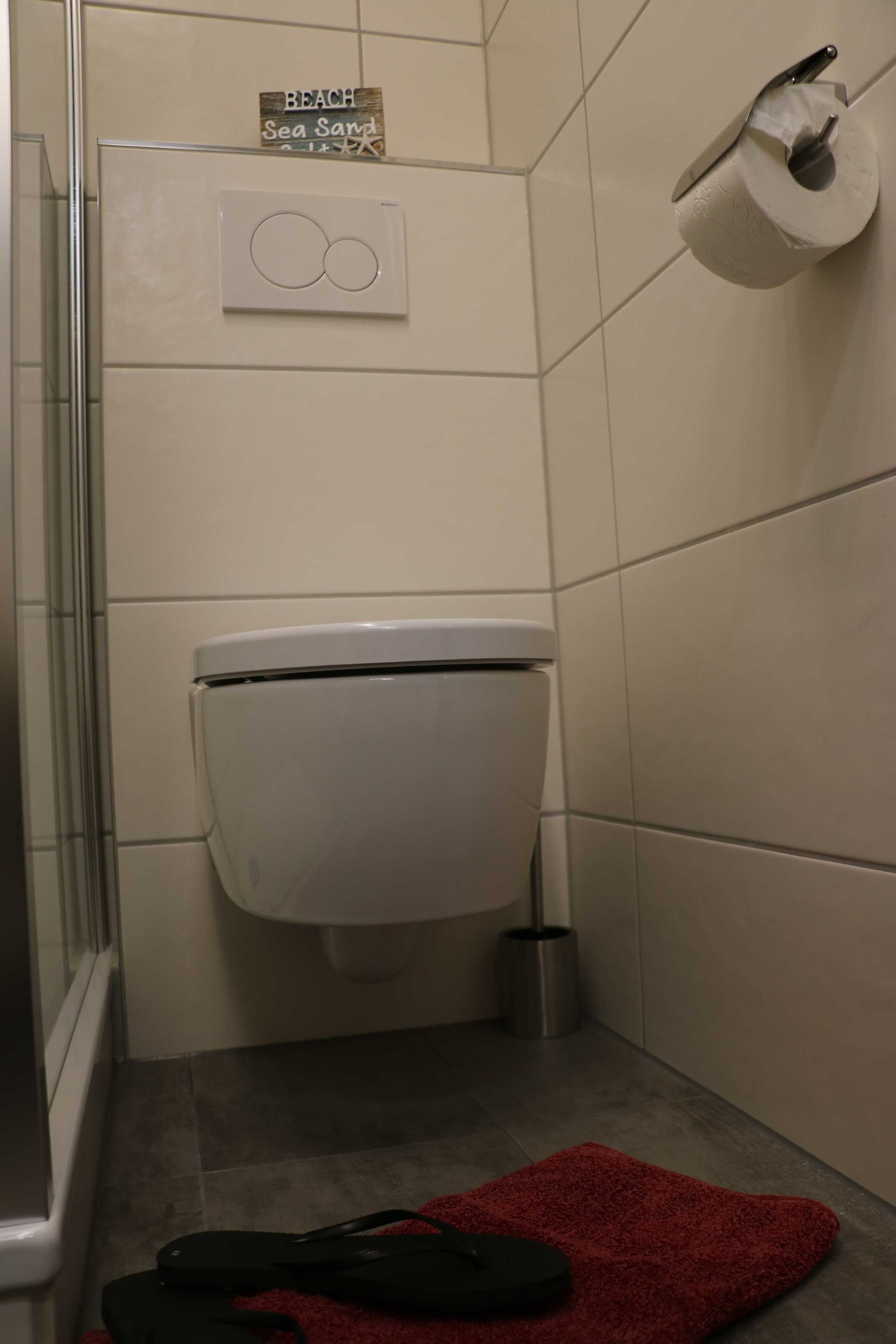 A bathroom with a toilet, a bidet, a red bath mat, and a 'BEACH Sea Sand' sign.