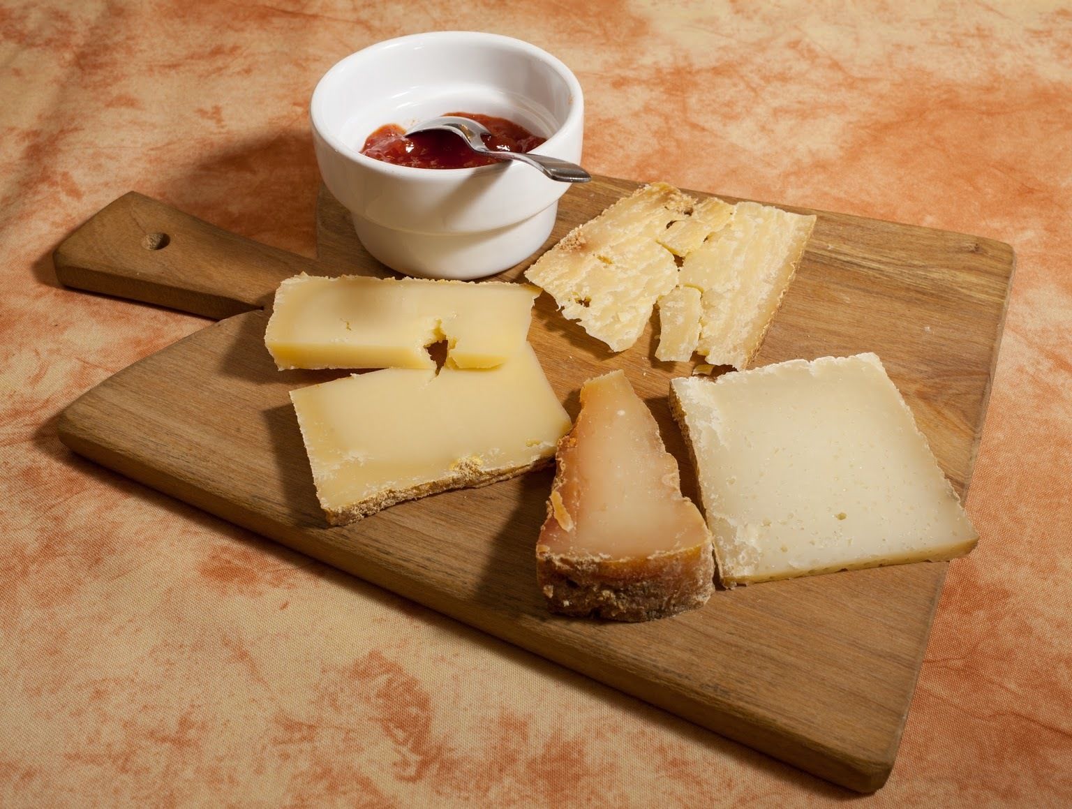 A wooden board with various cheese slices and a small bowl of jam.