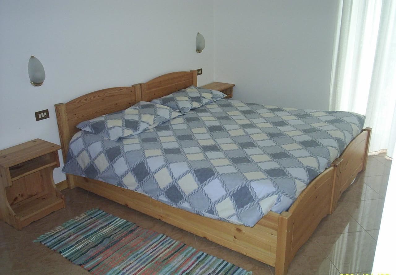 Una cama de madera con colcha a cuadros, una mesita de noche, un pequeño armario de madera y un tapete a rayas.
