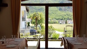 Un coin repas avec des tables préparées pour deux personnes, un banc et une vue sur un paysage verdoyant, visible à travers une grande porte vitrée.