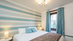 A hotel room with a striped wall, a bed with a brown blanket, a round nightstand with a lamp, and a window with blue curtains. - Springfield View (North Berwick)